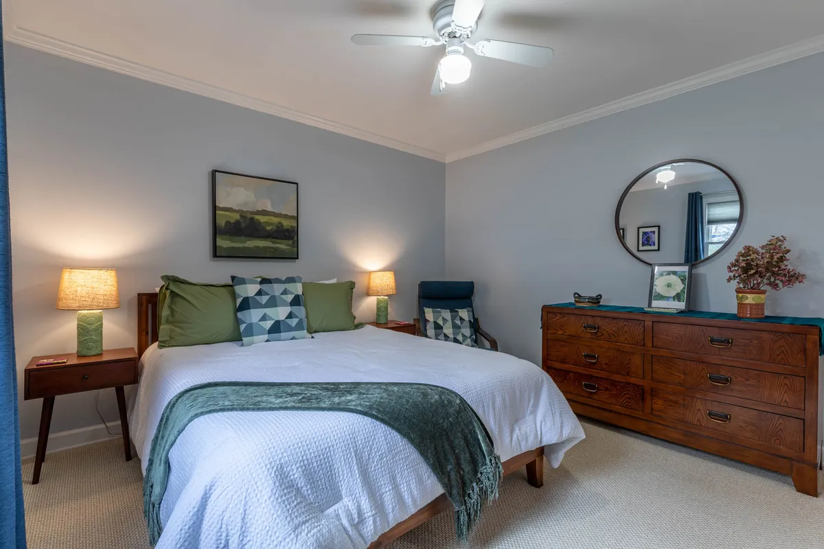 A serene bedroom with a bed dressed in green and blue accent pillows, a wooden dresser, soothing artwork, and table lamps that create a tranquil, cozy mood. The room is bathed in soft natural light, reinforcing the atmosphere of quiet retreat and relaxation.