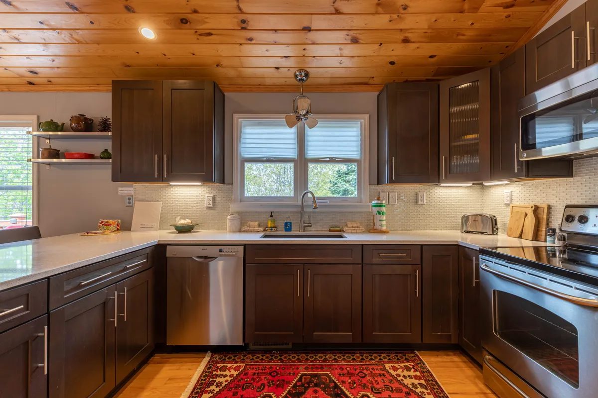 A cozy kitchen with a central island, stainless steel appliances, a wooden ceiling, and a colorful rug. The scene is warmly lit, emphasizing the practical layout and inviting ambiance, with ample countertop space and well-organized cabinetry, perfect for meal prep and gathering.