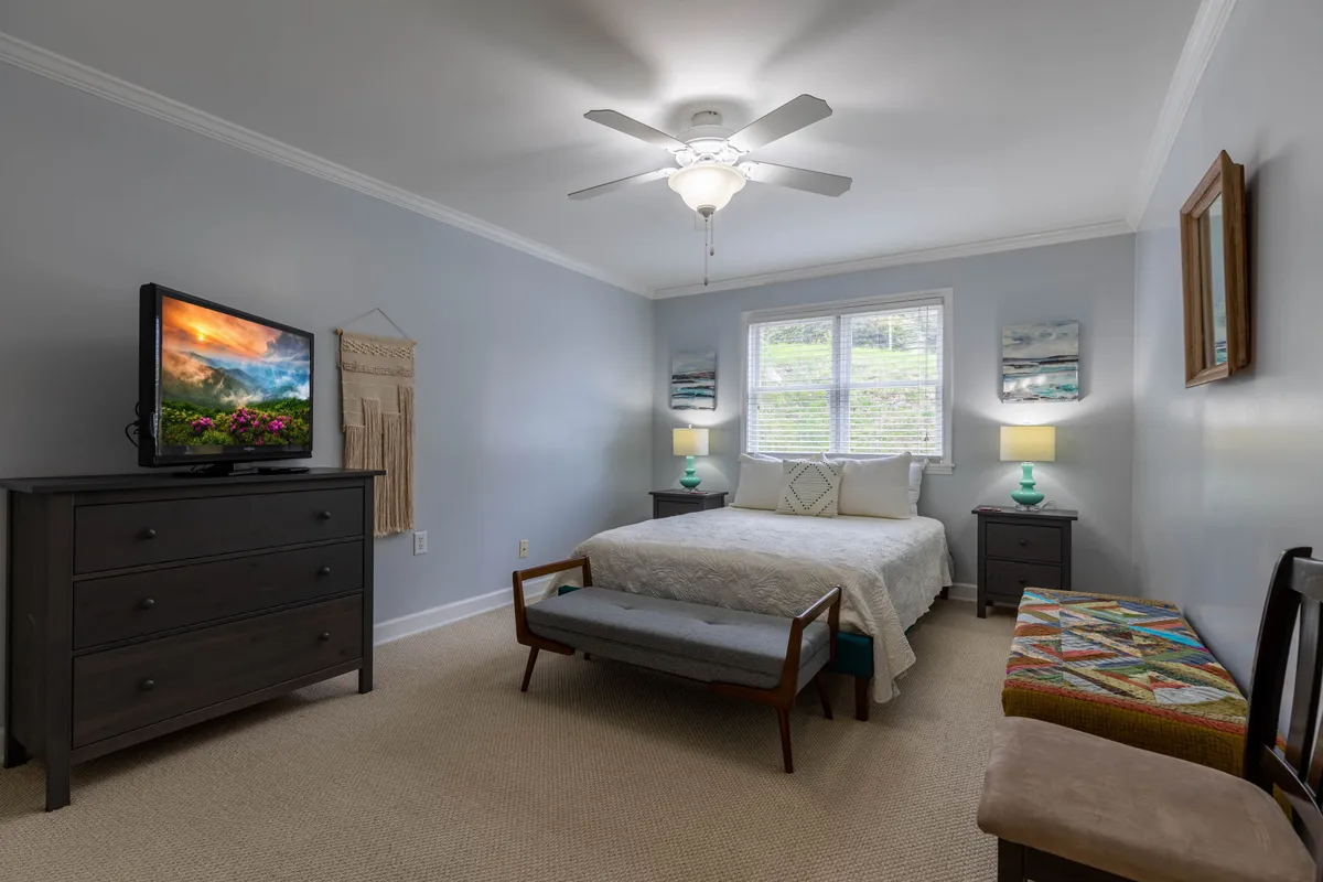 A cozy bedroom featuring a comfortable bed adorned with plush linens, bedside lamps providing a warm ambient glow, a bench at the foot of the bed, soft artwork above the headboard, and a television discreetly placed for relaxation. The room’s neutral tones and inviting textures evoke a sense of quiet comfort and privacy.