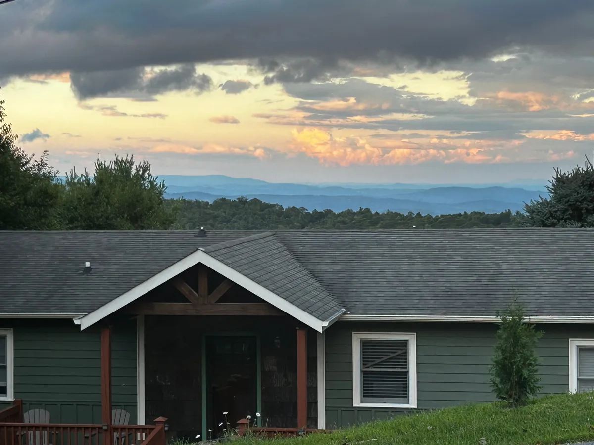 The house facade set against a backdrop of surrounding greenery and distant mountain ranges. A comfortable porch area invites relaxation, while the cloudy sky hints at the area's refreshing, high-altitude climate.