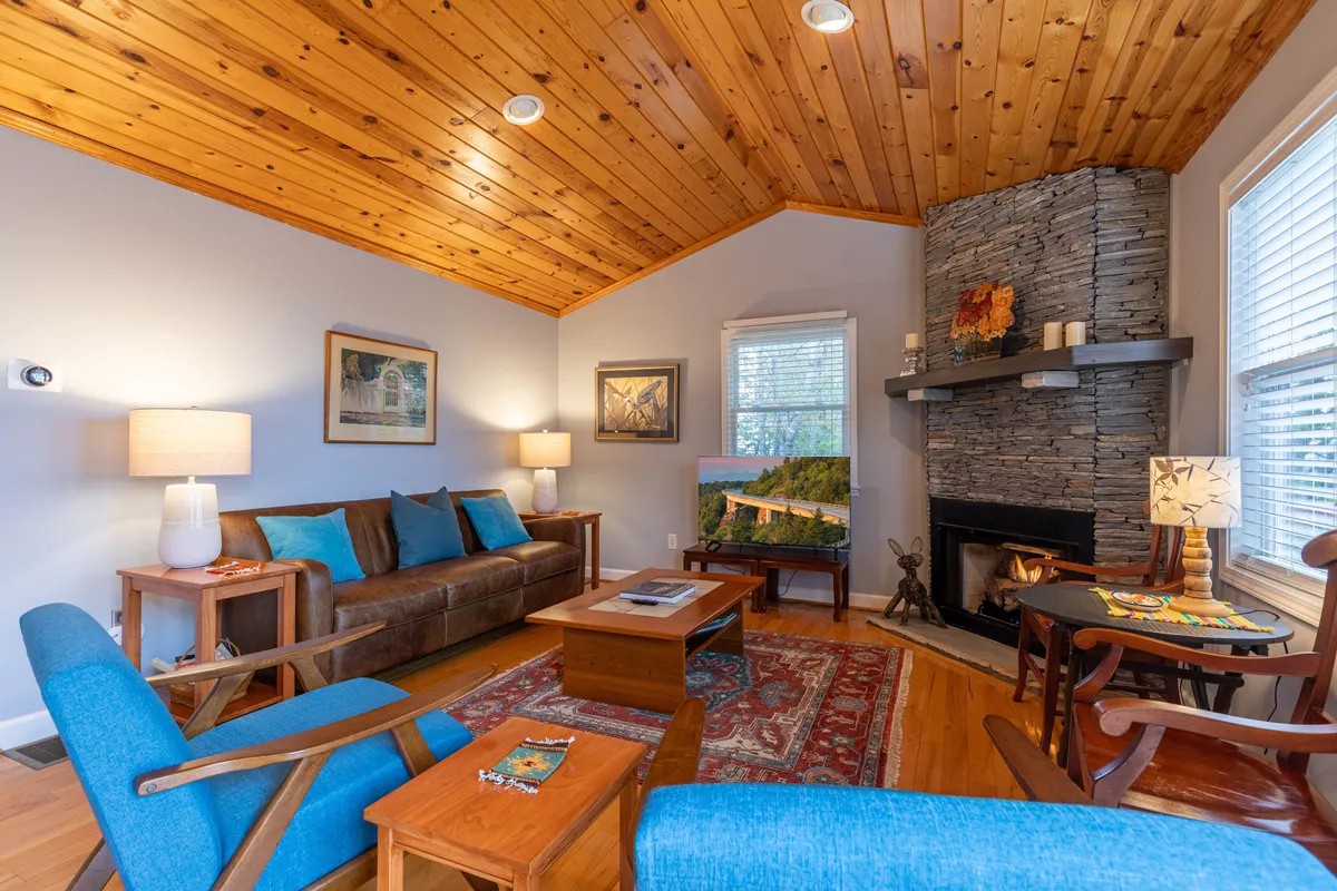 A warmly lit interior with a stone fireplace, wooden ceiling, tasteful wall art, and comfortable seating. The arrangement invites guests to relax, read, or socialize in a welcoming setting that blends rustic charm with modern comfort.