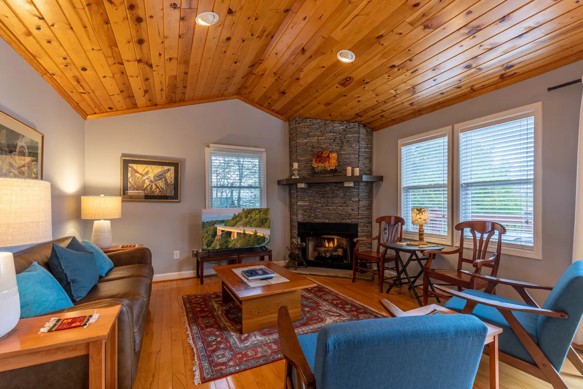 A cozy living room featuring a stone fireplace, wooden ceiling, and a comfortable sofa. Artwork adorns the walls, and large windows invite natural light, offering glimpses of the mountain scenery outside. The arrangement is inviting, with seating designed for relaxation and conversation.