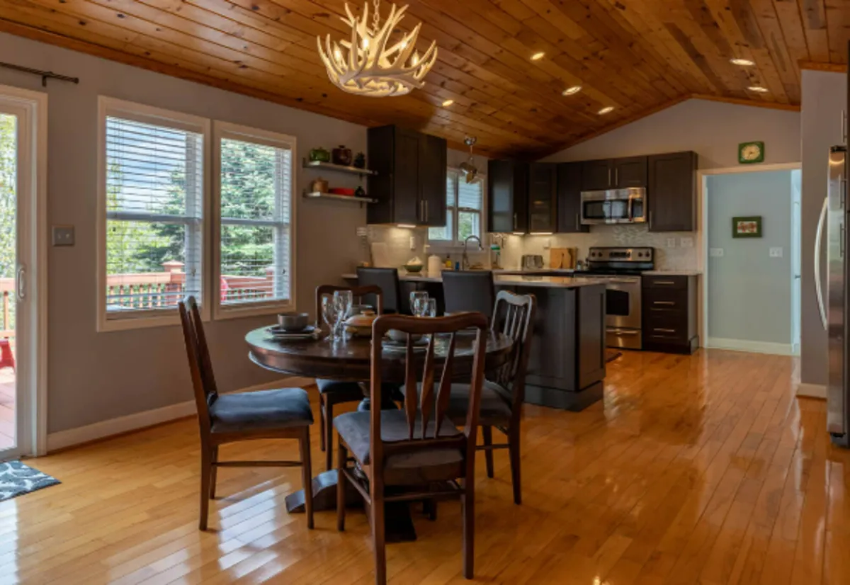 A combined kitchen and dining space with a dining table, kitchen island, large windows, and a chandelier overhead. The room is open and airy, allowing for natural light to flood in and providing glimpses of mountain vistas, reinforcing the seamless flow between kitchen and dining areas.