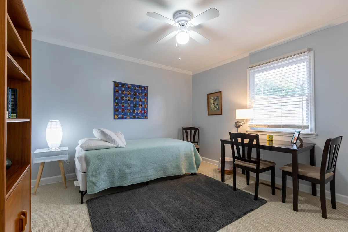 A bright interior space featuring a comfortable bed with a light green blanket, a wooden desk and chairs ideal for reading or remote work, colorful wall art, and a table lamp. The room is filled with natural light, emphasizing both relaxation and versatility.