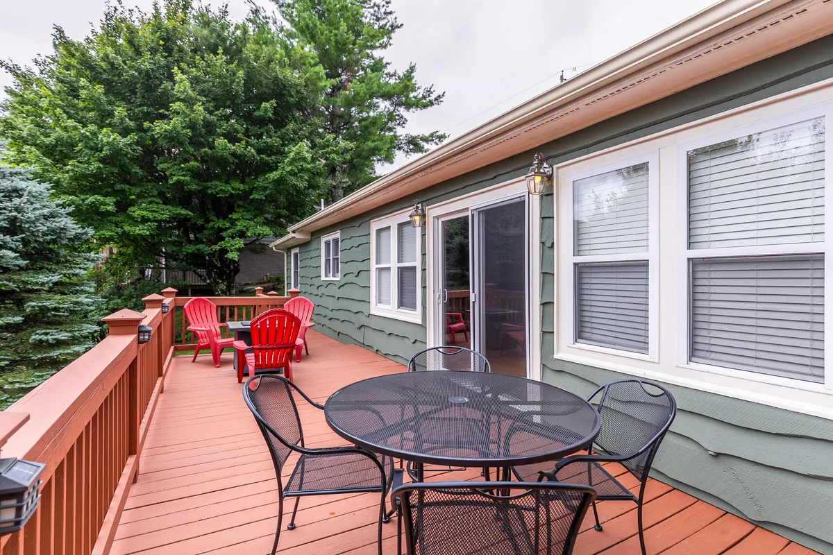 A cozy deck area with red Adirondack chairs and a black metal table, set against the green siding of the house and large shade trees in the background. The setup suggests a welcoming space for morning coffee, outdoor gatherings, or simply soaking in the mountain air.
