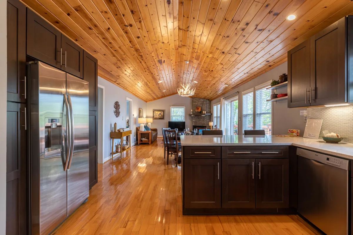 The living area boasts a wooden ceiling, kitchen island, dining area, large windows, and a striking stone fireplace. The space is filled with natural light and provides panoramic views of the mountains beyond, making it ideal for family gatherings or quiet moments alike.