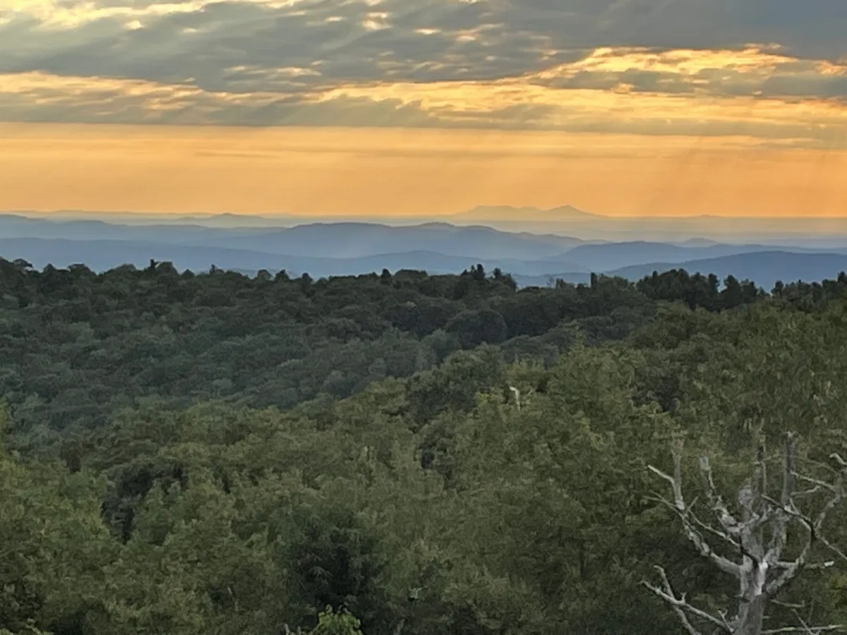 A breathtaking panoramic view of the Blue Ridge Mountains at sunset, with the vibrant sky casting a warm glow over layers of distant peaks and lush treetops. The tranquil scene is perfect for hiking, photography, or simply meditating in nature.