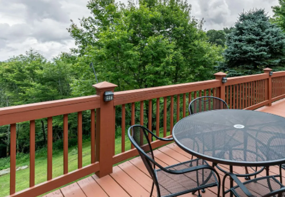This image captures the rear deck with its inviting circular table and black chairs, surrounded by lush green trees. The deck railing frames the outdoor scene, while the ambiance suggests a setting perfect for dining, relaxing, or enjoying panoramic views at sunrise or sunset.