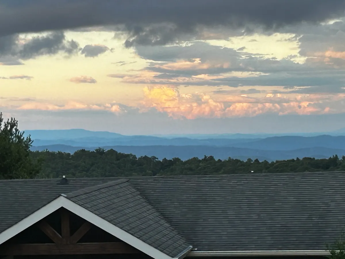 A panoramic view of a tranquil mountain range under a colorful sky, with the roofline of the cabin visible in the foreground. This scene suggests relaxing on the porch, stargazing, and enjoying the peacefulness of the mountain environment.